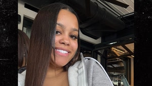A woman wearing a low-cut top and a gray zip-up hoodie is smiling while posing for a photo in what appears to be a home gym.