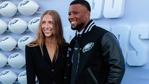 Saquon Barkley is seen with his fiancee Anna Congdon at an event for the Philadelphia Eagles. He's wearing a varsity jacket with the Eagles' logo on the front, while she's wearing a black jacket. There are several white footballs with the Eagles' logo set up behind them.