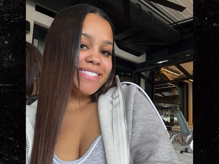 A woman wearing a low-cut top and a gray zip-up hoodie is smiling while posing for a photo in what appears to be a home gym.