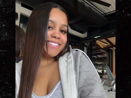 A woman wearing a low-cut top and a gray zip-up hoodie is smiling while posing for a photo in what appears to be a home gym.
