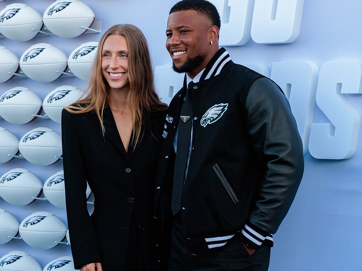 Saquon Barkley is seen with his fiancee Anna Congdon at an event for the Philadelphia Eagles. He's wearing a varsity jacket with the Eagles' logo on the front, while she's wearing a black jacket. There are several white footballs with the Eagles' logo set up behind them.