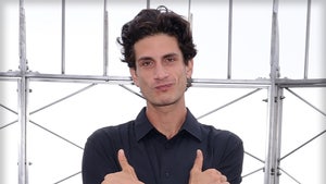 Jack Schlossberg is standing in front of several metal bars. His arms are folded across his chest, and he's sticking out his thumb and little fingers.