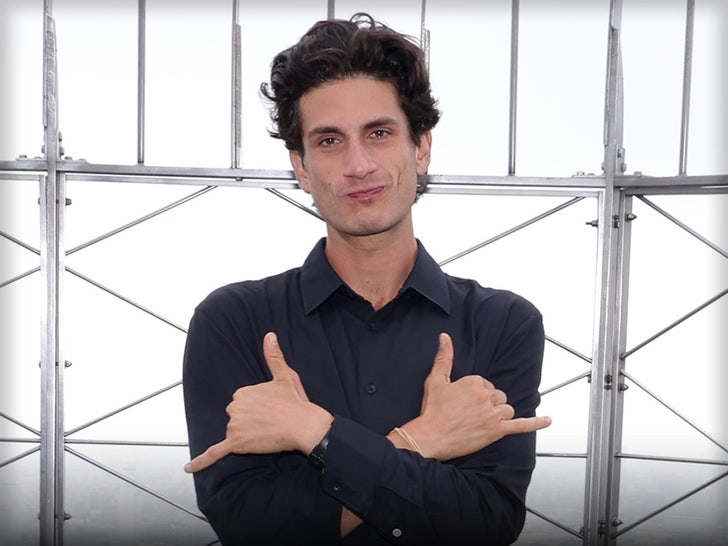 Jack Schlossberg is standing in front of several metal bars. His arms are folded across his chest, and he's sticking out his thumb and little fingers.