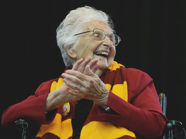 Sister Jean is seen clapping at a sports event. She's wearing a red long-sleeve shirt and a striped scarf.