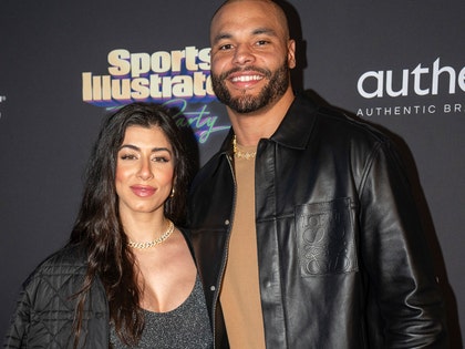Dak Prescott's standing next to his fiancee, Sarah Jane Ramos, at an event for Sports Illustrated. He's wearing a leather jacket and light brown shirt, while she's wearing a patterned jacket and a low-cut dress.