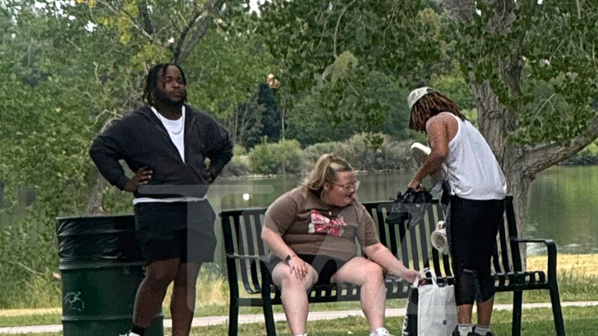 The image shows three people in a park near a body of water, with trees and greenery in the background. One person, wearing a brown t-shirt and shorts, is sitting on a black metal bench and reaching into a bag on the ground. Another person, dressed in a white tank top, black pants, and a backward white baseball cap, is standing next to the bench, holding items in their hands. The third person, wearing a black jacket and shorts, stands nearby with hands on their hips, looking ahead. A green trash bin is visible beside the standing person in the jacket.