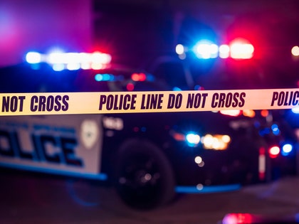 A pair of police cars with flashing red and blue lights are parked behind a line of police tape with text reading 