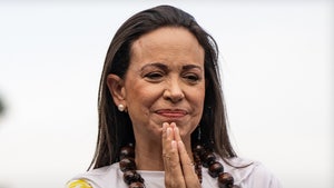 Maria Corina Machado is seen placing her hands together. She's wearing a multicolored t-shirt and several necklaces.