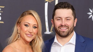 Emily Wilkinson and Baker Mayfield are standing next to each other at an event. She's wearing a brown dress, while he opted for a white button-up shirt and a dark blue jacket.