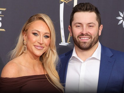 Emily Wilkinson and Baker Mayfield are standing next to each other at an event. She's wearing a brown dress, while he opted for a white button-up shirt and a dark blue jacket.