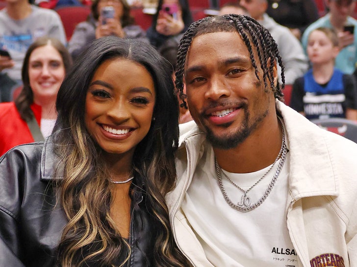 Simone Biles and her husband Jonathan Owens are smiling while sitting next to each other. She's wearing a leather jacket, and he's dressed in a white t-shirt underneath a white jacket.