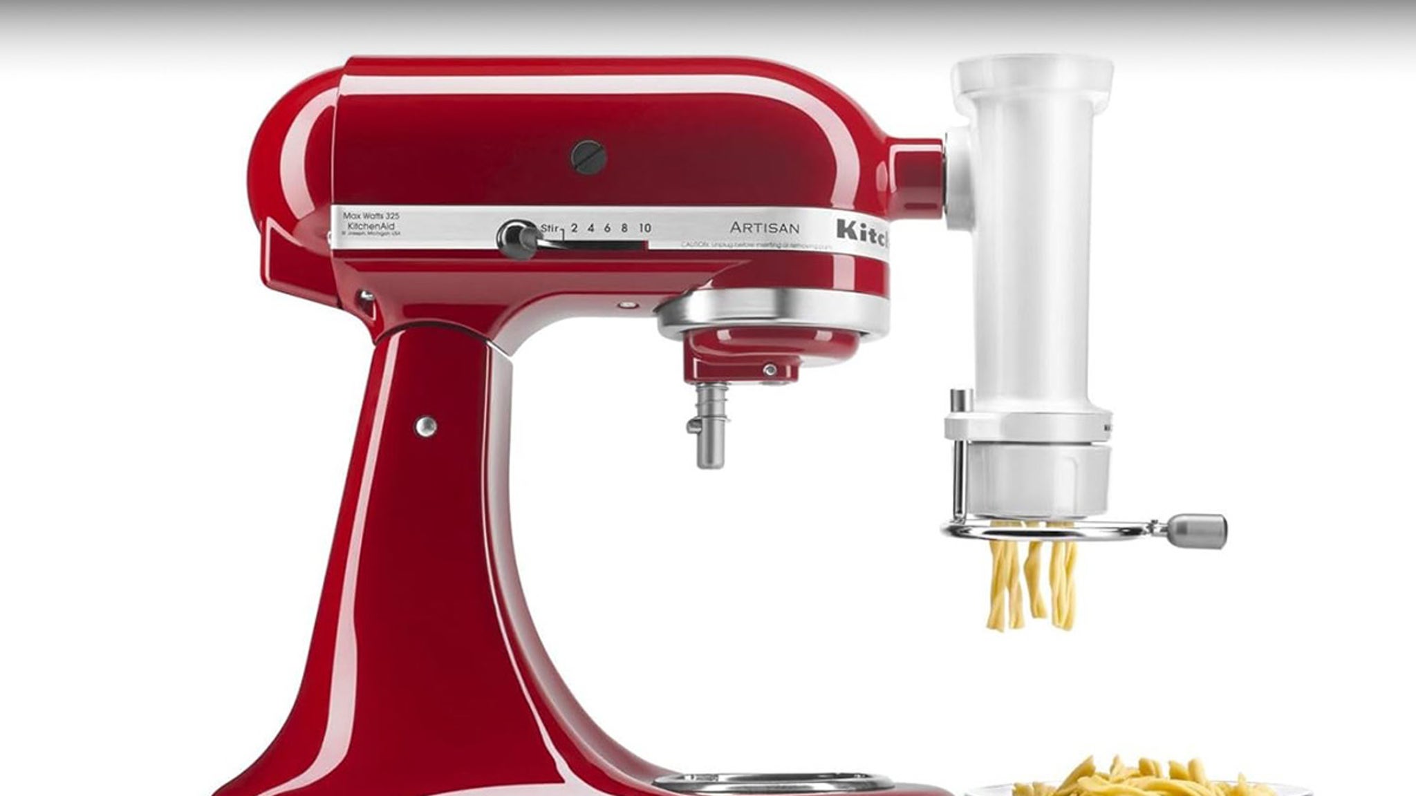 The image shows a close-up of a red KitchenAid stand mixer with a white pasta-making attachment. The mixer has a sleek, glossy finish with a silver control lever marked with numbers for different speeds on the side. The pasta attachment is extruding twisted pasta strands onto a white plate placed below it. The setup is displayed against a plain white background.