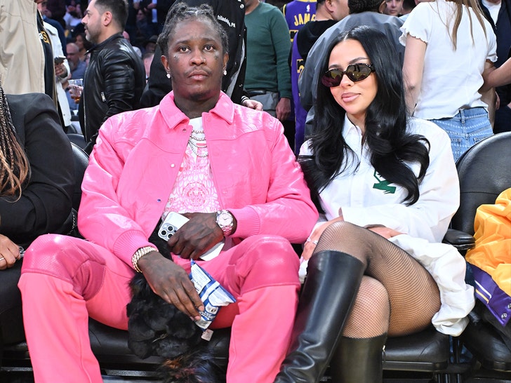 Mariah The Scientist and Young Thug are seen posing for a photo at a basketball game. He's wearing an all-pink outfit, while she's wearing a white blouse, fishnet leggings, leather boots and dark sunglasses.