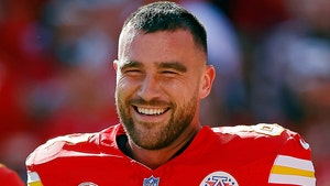 A smiling man is ween wearing a red football jersey with yellow-and-white numbering and decals. The jersey also features several patches on its front portion.