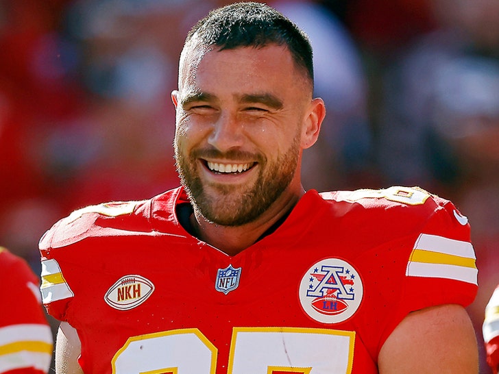 A smiling man is ween wearing a red football jersey with yellow-and-white numbering and decals. The jersey also features several patches on its front portion.