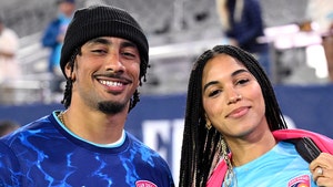 Jordan Love is smiling while posing for a photo next to his wife Ronika. They're both wearing San Diego Wave FC jerseys, his is dark blue and hers is much lighter. She's also got a pink scarf.