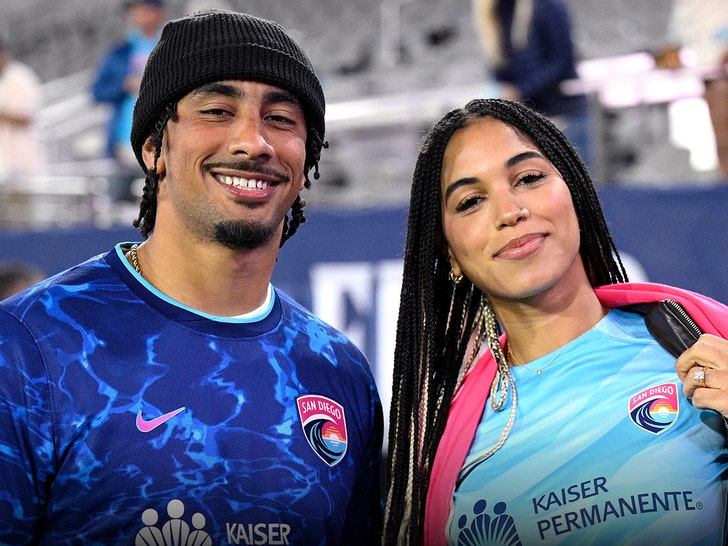 Jordan Love is smiling while posing for a photo next to his wife Ronika. They're both wearing San Diego Wave FC jerseys, his is dark blue and hers is much lighter. She's also got a pink scarf.