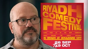 David Cross is looking concerned next to a poster for the Riyadh Comedy Festival. He's wearing a gray button-up shirt.