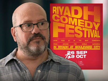 David Cross is looking concerned next to a poster for the Riyadh Comedy Festival. He's wearing a gray button-up shirt.