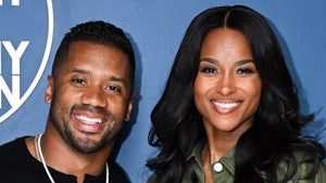 A man wearing a black shirt and a diamond necklace is seen posing next to a woman wearing a green button-up jacket in front of a blue surface.