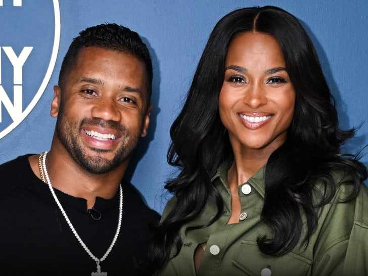 A man wearing a black shirt and a diamond necklace is seen posing next to a woman wearing a green button-up jacket in front of a blue surface.