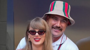 Travis Kelce is seen standing behind Taylor Swift. She's wearing a checkered dress, gold jewelry and sunglasses, and he's wearing a Gucci poli and a near-matching bucket hat.