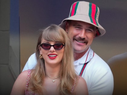 Travis Kelce is seen standing behind Taylor Swift. She's wearing a checkered dress, gold jewelry and sunglasses, and he's wearing a Gucci poli and a near-matching bucket hat.