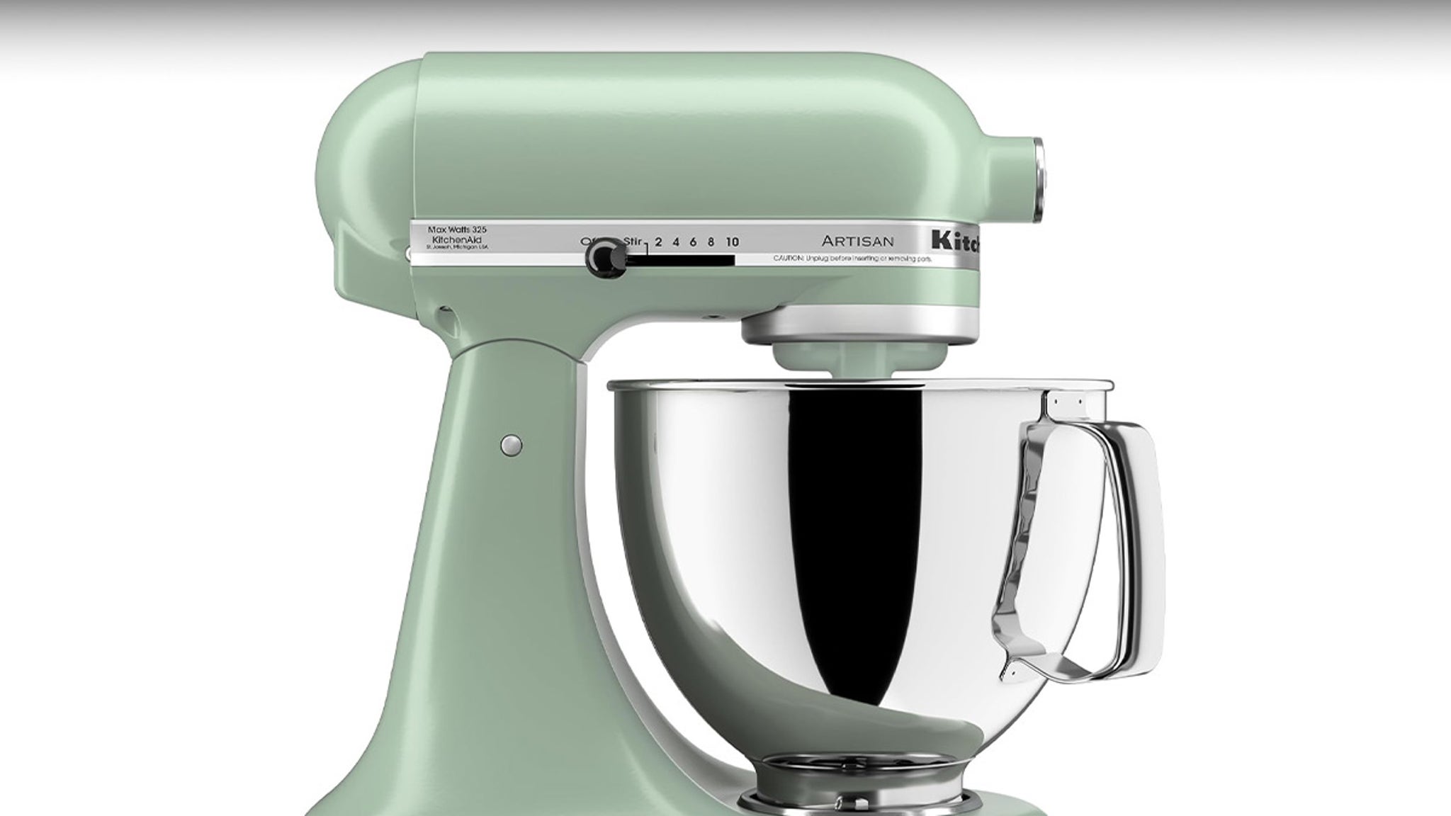 The image shows a stand mixer in a muted pastel green color with a smooth, rounded design. The mixer has a large, shiny stainless steel mixing bowl attached, featuring a sturdy handle on the right side. On the mixer's head, there is a speed control dial marked with numbers from 0 to 10, labeled 'Stir' and 'Artisan,' along with branding for KitchenAid. The overall look is sleek and modern, set against a plain white background.