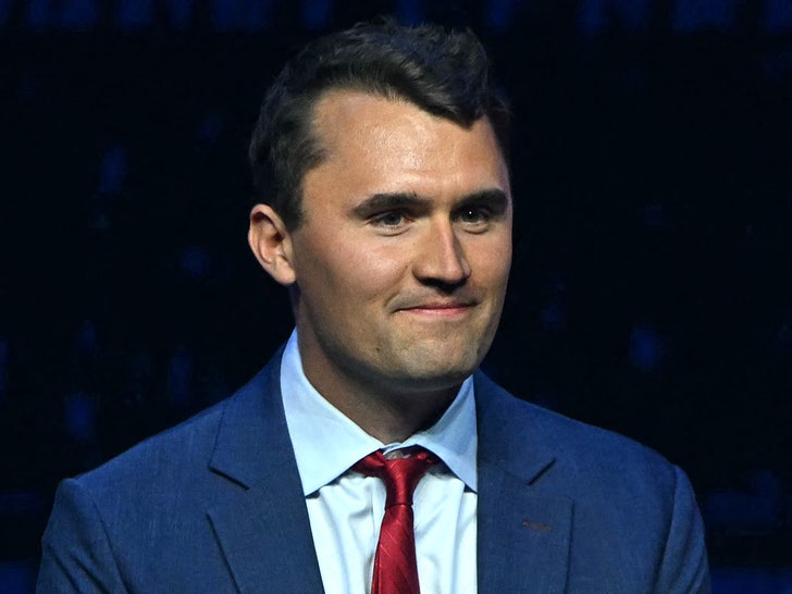 Charlie Kirk is seen while flashing a slight smile. He's wearing a dark blue jacket, a light blue shirt, and a red tie.