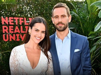 A woman wearing a white lace dress is standing next to a man wearing a light blue button-up shirt and a navy blue sports coat are standing in front of a hedge and various greenery. They're standing in front of a sign partially reading 